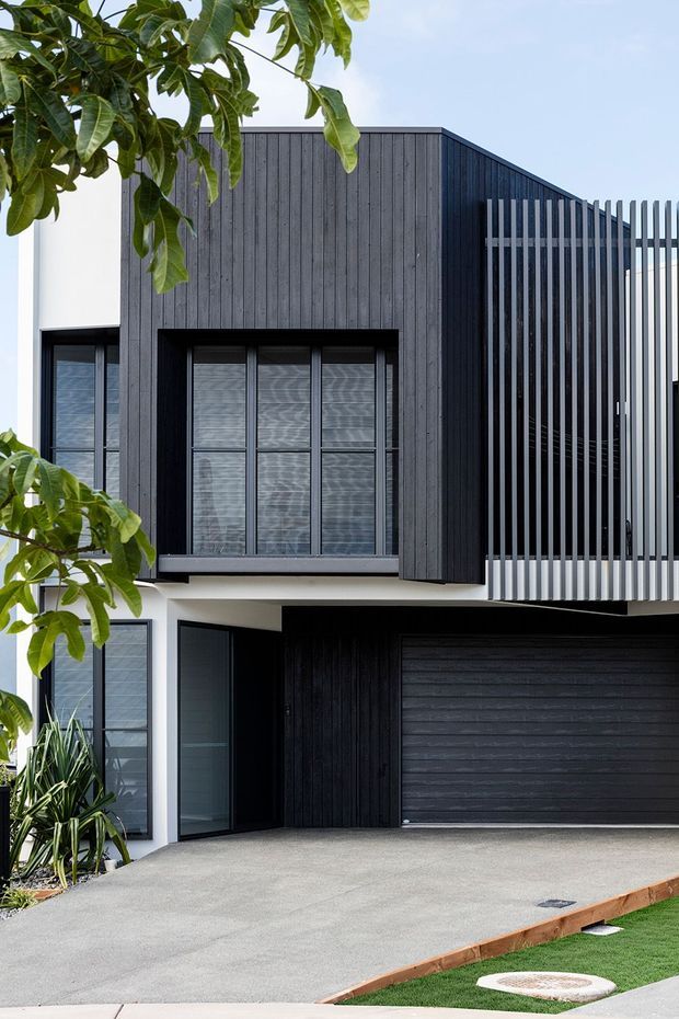 Modern two-storey residential facade with dark vertical timber cladding, white rendered walls, black-framed windows, a large dark garage door, and a prominent vertical slatted privacy screen. A concrete driveway and landscaped planting sit in front of the home.