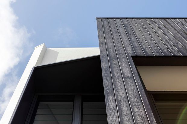 Upward-looking close view of a contemporary building facade against a blue sky, featuring dark vertical weathered timber cladding, black-framed louvred windows, crisp white rendered walls, and strong angular roof and wall lines.