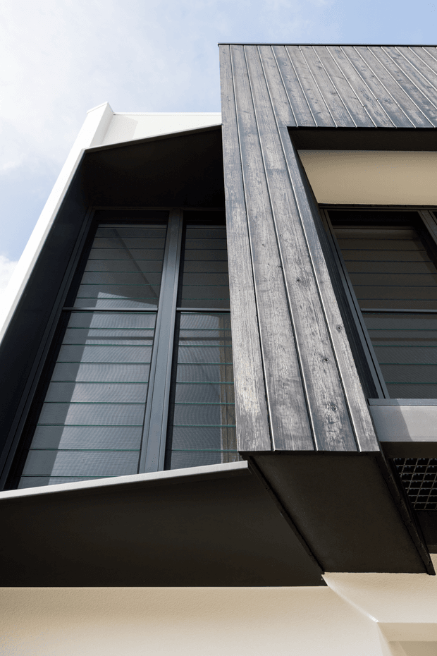 Upward-looking close-up of a contemporary building facade, featuring dark metal-framed louvered windows, projecting eaves, and a prominent vertical weathered timber-clad element. White rendered walls and roof edges contrast with the charcoal facade components against a pale sky.