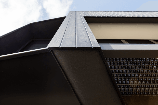 Upward-looking close-up of a contemporary building facade, showing dark angular cladding, a standing-seam metal roof or canopy, a deep overhang, and a patterned breeze-block screen beneath a recessed opening. The composition emphasizes strong geometric forms and contrasting light and dark surfaces.