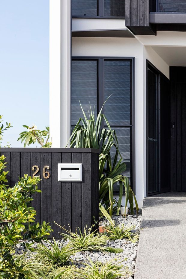 Bright exterior view of a contemporary residential entry. A white rendered facade with dark-framed windows and a recessed black front door sits behind a low vertical dark timber boundary wall marked with the house number 26 and a white letterbox. The entry path is bordered by grey gravel and drought-tolerant landscaped planting, with strong black-and-white contrast and clean geometric lines.