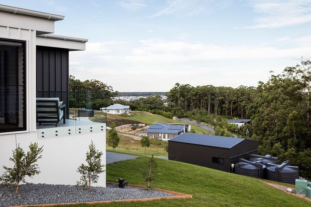 Exterior view of a contemporary multi-level residence on a sloping, semi-rural site, with a white rendered facade, dark vertical cladding, projecting flat roof forms, glass balcony balustrade, and broad views across surrounding trees and neighbouring homes. The landscaped foreground includes lawn, gravel beds, and young plantings.