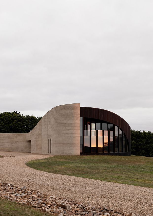 Exterior view of a contemporary curved building set within a landscaped rural-looking site. The composition combines a sculptural pale concrete wall with a dark curved roof and tall floor-to-ceiling glazed facade, reflecting the warm evening sky. A gravel driveway and rock-edged lawn occupy the foreground, with dense hedging behind the building.