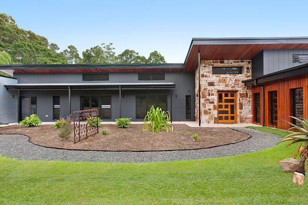 Single-storey contemporary house exterior with a low-pitched roof and broad eaves. The facade combines dark grey cladding, warm timber-lined soffits and entry surrounds, and a prominent natural stone feature wall around the timber-framed front door. A curved gravel path and landscaped garden lead to the entrance, with established trees in the background.