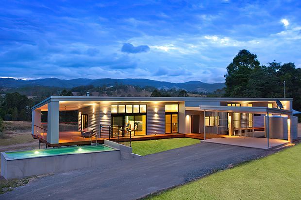 Twilight exterior view of a contemporary single-storey home with horizontal dark cladding, broad glazed openings, flat roof forms, covered outdoor living areas, a driveway, and a raised rectangular swimming pool. Rolling wooded hills form the distant background.