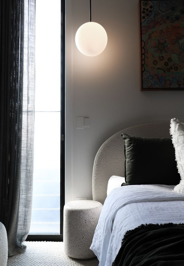 A close-up bedroom interior featuring a textured curved upholstered headboard, white bedding, black and white pillows, and a black throw. A softly glowing spherical pendant hangs beside a tall window dressed with sheer dark curtains. The room has a calm, monochromatic palette with tactile finishes and abstract artwork above the bed.