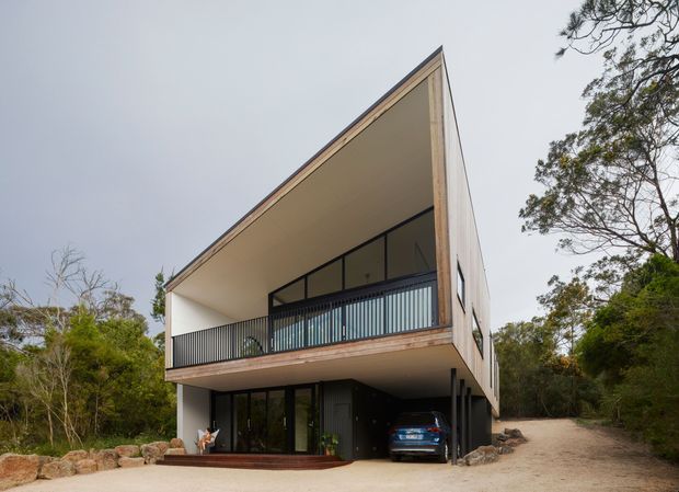 Exterior view of a contemporary two-storey house set among trees, featuring a dramatic steeply sloping roof plane, large open upper-level balcony with black metal balustrades, expansive glazing, pale rendered walls, and timber-toned trim. A vehicle is parked beneath the upper floor in an integrated ground-level carport.