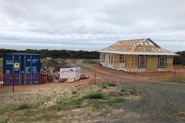 A single-storey residential house under construction on a coastal-looking site. The timber-framed walls and exposed trussed roof are visible, with installed windows and doors, temporary orange safety fencing, a shipping container, building materials, and an unsealed gravel access area. Overcast sky and low coastal vegetation form the background.