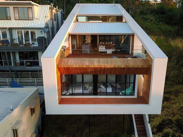 Exterior elevated view of a narrow multi-storey contemporary residence on a sloping, densely vegetated site. The house has a distinctive large white gabled frame enclosing stacked glazed living areas and timber-lined balconies, with a corrugated metal pitched roof, extensive black-framed glazing, and an external stair descending along one side. Adjacent residential buildings are visible closely beside it.