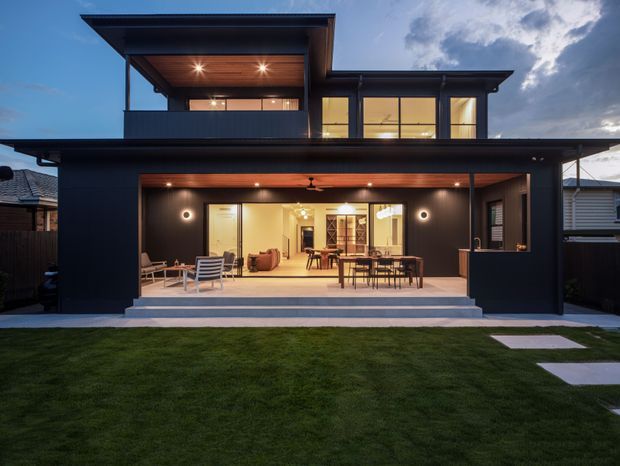 A contemporary two-storey residential house viewed from the rear garden at dusk. The dark-clad facade features broad floor-to-ceiling glazing, a deep covered outdoor entertaining area with timber-lined ceiling, an upper-level balcony or recessed terrace, and layered flat roof forms. Interior living and dining areas are visible through the open glazing, with a lawn and stepping-stone path in the foreground.