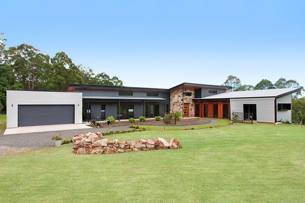 Single-storey contemporary residential house viewed from the front across a broad lawn. The building has a low, angular roofline, white horizontal cladding, dark-framed glazing, a large integrated garage, and a central feature wall of irregular natural stone with warm timber-toned entry elements. A gravel driveway, landscaped garden, and low stone fire-pit or retaining feature occupy the foreground.