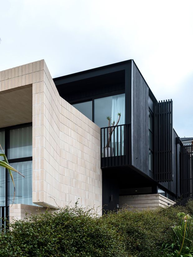 Exterior architectural photograph of a contemporary multi-level residence, featuring a strongly geometric composition of pale masonry walls and projecting volumes contrasted with dark vertical cladding, black-framed glazing, recessed balconies, and vertical privacy screens. Dense planting partially obscures the lower facade.