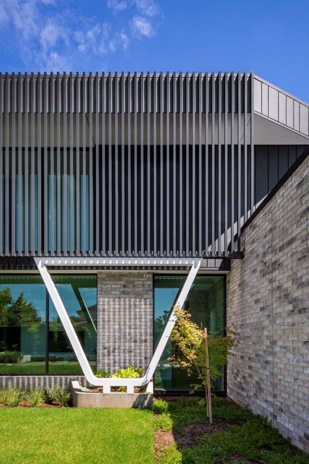 Exterior architectural photograph of a contemporary multi-level building with a strong vertical metal louvre screen across the upper facade, large dark-framed glazed openings at ground level, and a pale grey masonry wall. A distinctive white V-shaped structural or sculptural element projects in front of the glazing. The image is set against a bright blue sky with a manicured lawn and planted garden bed in the foreground.