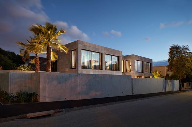Street-level exterior view of a modern two-storey residence at dusk, enclosed by a tall smooth concrete boundary wall. The house has strongly geometric cubic volumes, large floor-to-ceiling windows, dark metal window frames, and a flat roof. Palm trees and landscaping are visible within the property, with warm interior lighting contrasting against the cool blue evening sky.
