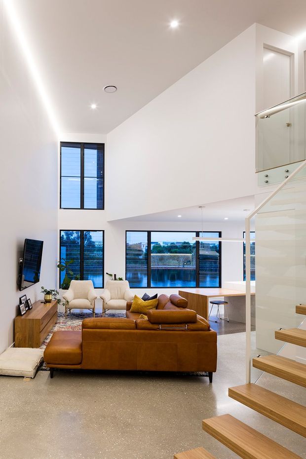 Bright double-height contemporary living room viewed from an open stairway, featuring a large caramel leather sectional sofa, polished concrete or terrazzo-like flooring, white walls, black-framed glazing, and a floating timber stair with a glass balustrade. A compact kitchen or dining counter is visible beyond the seating area, with water and buildings seen through the rear windows.