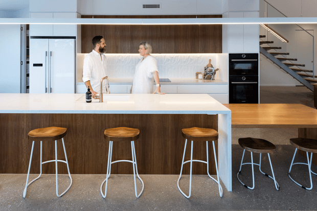 Contemporary open-plan kitchen with a large white island and waterfall edge, dark timber cabinetry, integrated appliances, and a softly veined light stone backsplash. Timber bar stools line the island, while a staircase and timber flooring are visible in the adjoining space. The kitchen is brightly lit and occupied by two people.