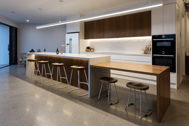 A contemporary open-plan kitchen dominated by a large white island with integrated seating, timber cabinetry, and a timber breakfast table extension. The kitchen includes white lower cabinetry, dark timber overhead cabinetry, integrated black ovens, a textured white feature splashback, and warm under-cabinet lighting. Six slim bar stools line the island and two additional stools sit at the timber dining extension. A polished grey concrete floor, minimalist linear pendant light, and expansive glazing contribute to the refined modern aesthetic.