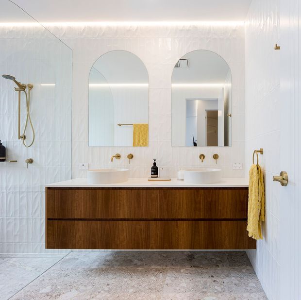 Bright contemporary bathroom featuring a floating dark timber double vanity with two white vessel basins, brass wall-mounted taps, arched mirrors, white textured wall tiles, a walk-in shower, and pale stone-look flooring. Yellow towels provide a small accent of colour.