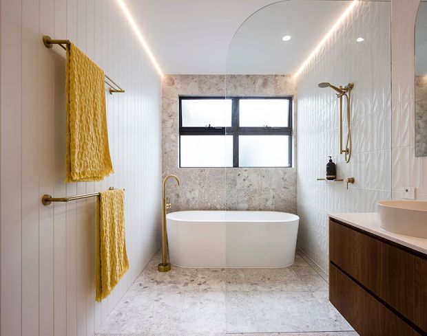 Bright contemporary bathroom featuring a freestanding white bathtub beneath a black-framed window, a separate brass shower, white vertically ribbed wall tiles, pale stone-look floor and wall tiles, and dark timber vanity cabinetry. Mustard-yellow towels and brass fixtures add warm accents.