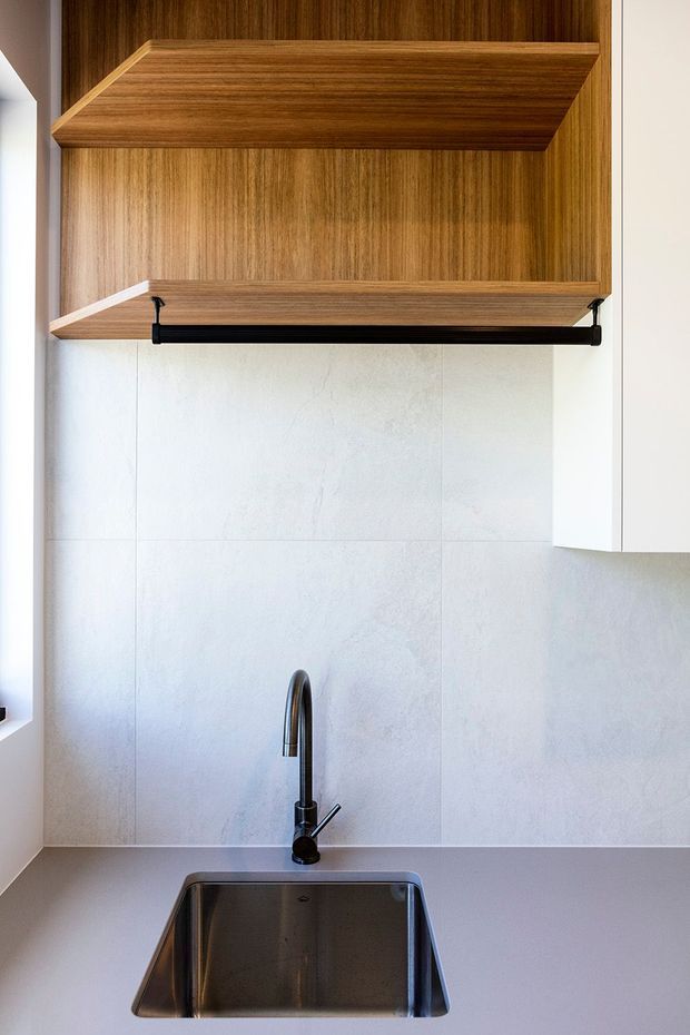 Close-up view of a compact contemporary kitchen sink area, featuring a stainless-steel undermount sink, dark curved tapware, pale stone or porcelain splashback, white cabinetry, and warm wood open shelving with an integrated hanging rail.