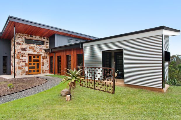 Exterior view of a contemporary single-storey home with a low-pitched roof, bold horizontal metal cladding, warm timber soffits and wall panels, and a distinctive natural stone entry facade. The home sits within a landscaped garden with gravel paths and a decorative patterned privacy screen.