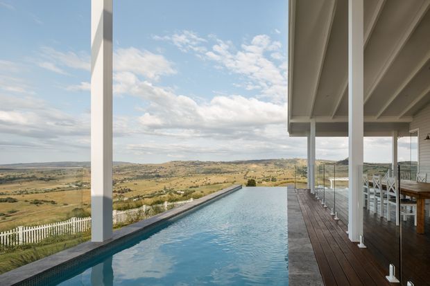 A modern elevated residence overlooking a broad rural landscape, featuring a long infinity-edge swimming pool beside a timber deck and covered outdoor entertaining area. Slim white structural columns, glass balustrades, and expansive open views create a bright, contemporary indoor-outdoor setting.