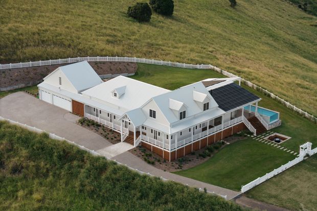 Aerial view of a large, light-coloured rural residence with multiple connected gabled roof forms, wraparound verandas, white picket fencing, landscaped lawns, a paved driveway, and a prominent rooftop solar-panel array. The property sits within open grassy countryside.
