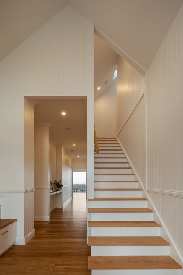 A bright residential stairway and hallway interior with an open-riser staircase featuring warm timber treads and white painted risers. The space has white vertical wall panelling, detailed skirting and dado-style trim, recessed lighting, timber flooring, and a high sloping ceiling. A glazed opening at the end of the hall provides a distant view outside.