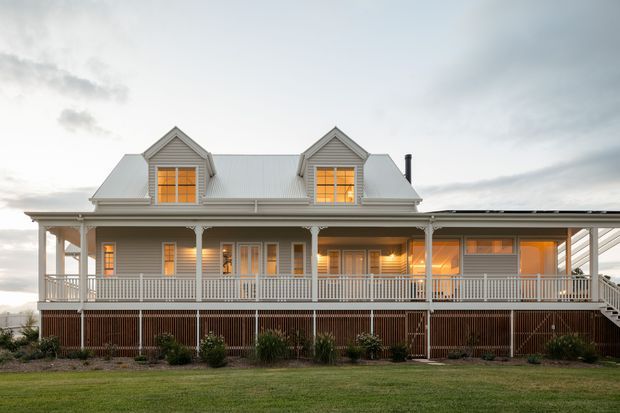 Exterior view of a raised, symmetrical weatherboard house with a broad covered verandah, twin dormer windows, a pitched metal roof, white posts and railings, and warm interior light glowing through the windows. The home sits above a landscaped lower level with timber screening and a lawn in the foreground.