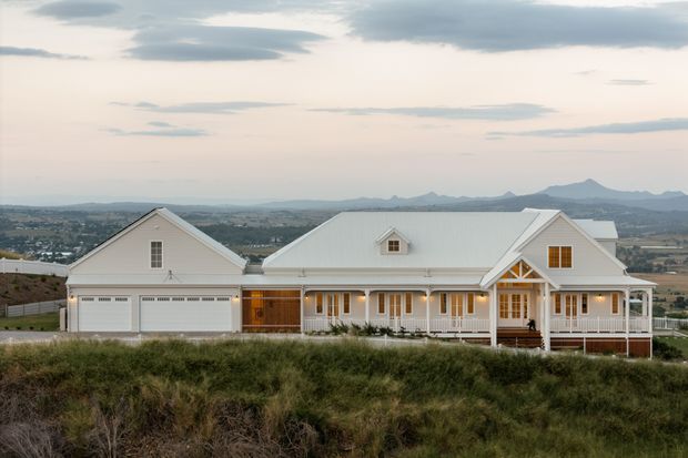 A wide exterior photograph of a large, symmetrical white farmhouse-style residence on an elevated site. The single-storey home has multiple gabled roof sections, a light metal roof, covered front verandah, white timber detailing, dormer windows, and an attached double garage. A broad rural landscape and distant mountains are visible behind the property.