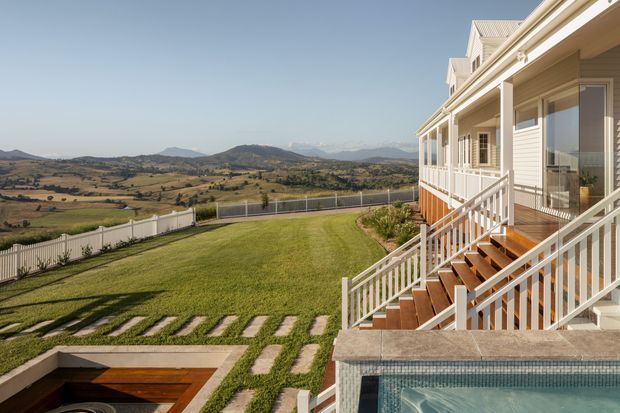 A multi-level light-coloured house exterior overlooking a broad rural valley and rolling hills. The facade features horizontal weatherboard cladding, repeated gabled roof forms, white decorative trim and balustrades, and timber staircases leading down to a landscaped lawn. A tiled swimming pool and built-in outdoor area are visible in the foreground.