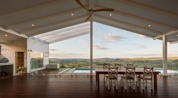A spacious open-plan indoor-outdoor living and dining area with a timber-lined ceiling, exposed beams, recessed lighting, large glass openings, and a broad timber deck. A dining table faces a rectangular swimming pool and expansive rolling landscape views. The room also includes a fireplace, built-in cabinetry, a ceiling fan, and a partially enclosed outdoor area.