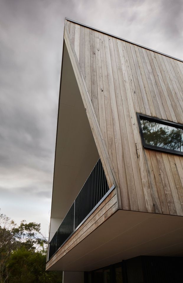 Upward-looking exterior view of a contemporary multi-level building with pale vertical timber cladding, sharp angular roof and wall geometry, deep cantilevered overhangs, dark metal balcony balustrades, and black-framed horizontal glazing. The overcast sky and surrounding trees frame the facade.