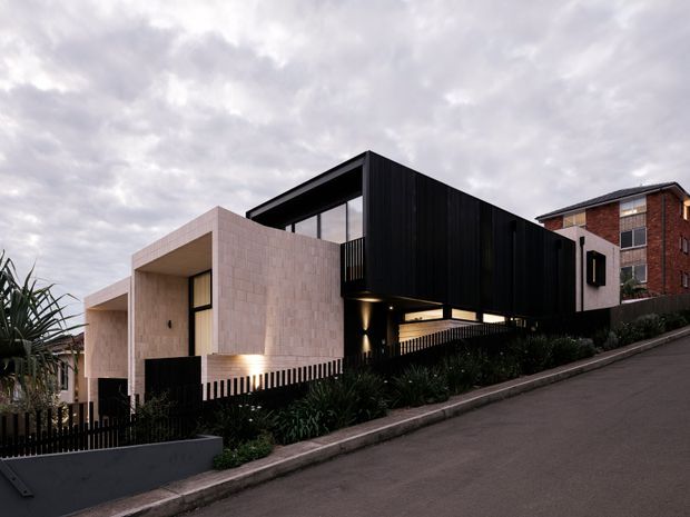 Exterior view of a contemporary multi-level residence on a sloping suburban street. The home combines pale masonry volumes with a dramatic black upper box, extensive glazing, dark vertical cladding, balconies and integrated exterior lighting. Landscaping and a dark metal fence line the street frontage, with a neighbouring brick dwelling visible behind.