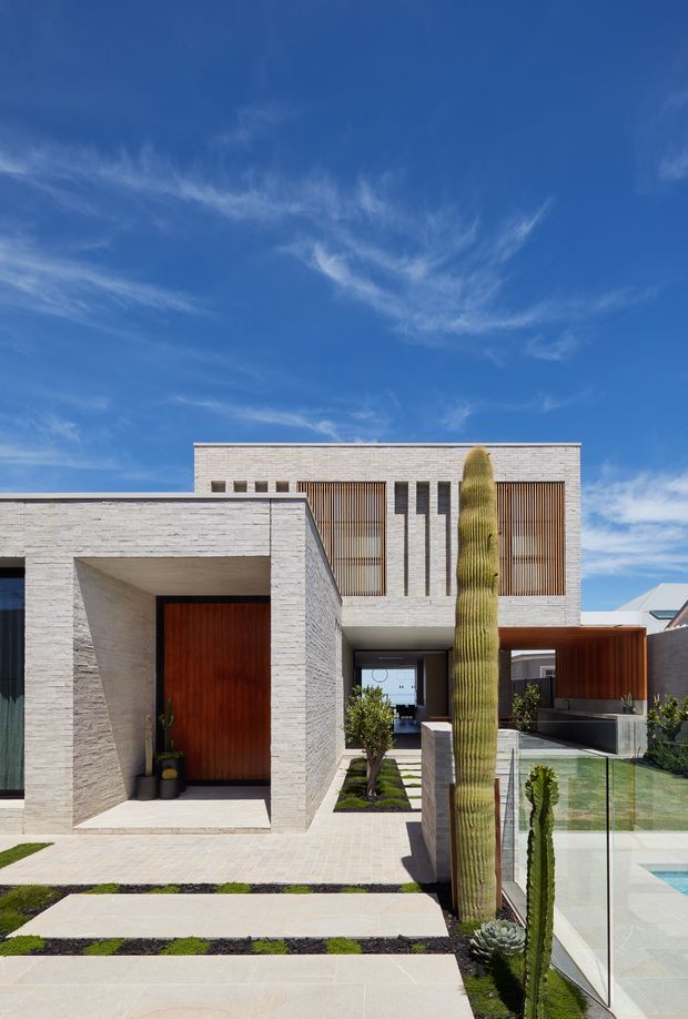 Contemporary multi-level residential facade photographed from the front, featuring pale horizontal masonry, strong rectilinear volumes, timber entry and screening elements, and extensive glazing. A landscaped forecourt with sculptural columnar cacti, paved stepping paths, low planting, and a glass-edged pool or water feature occupies the foreground under a bright blue sky.