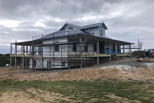 An elevated two-storey house under construction, surrounded by scaffolding. The building has a dark pitched roof with multiple gables, a wraparound verandah, light exterior walls, and several dormer-like upper windows. The site is unfinished with exposed framing, construction materials, and earthworks under an overcast sky.