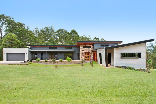 A wide exterior view of a contemporary single-storey residence set behind a broad lawn and surrounded by mature trees. The house features low-pitched intersecting roof forms, white horizontal cladding, dark-framed glazing, a projecting garage, and a central entry marked by a distinctive timber and glazed screen.