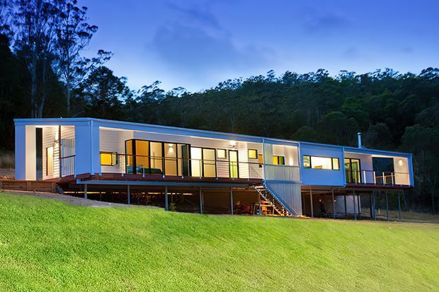 A long, elevated single-storey modern home set on a sloping grassy site at dusk, surrounded by dense bushland. The building features pale blue-grey horizontal cladding, extensive illuminated glazing, a raised deck and open undercroft supported by slender posts, with an exterior stair leading to the deck.