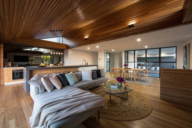 Open-plan contemporary living, kitchen and dining area with a large grey sectional sofa in the foreground, a timber kitchen island and integrated cabinetry, timber flooring, and extensive warm timber slatted ceiling lining. The dining area sits beside broad black-framed windows with horizontal blinds, while recessed and pendant lighting create a warm, inviting atmosphere.