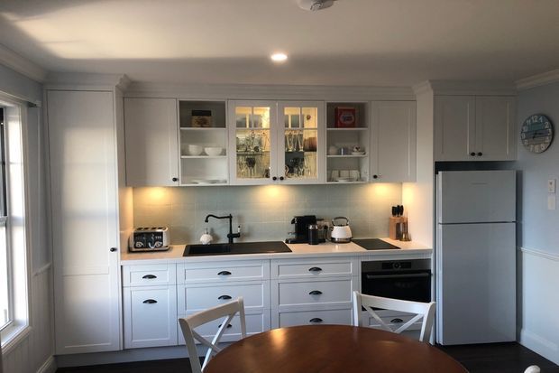 A compact, traditional-style kitchen and dining area with white shaker cabinetry, glass-front upper cupboards, pale tiled backsplash, dark benchtop, stainless-steel refrigerator and built-in oven. A round timber dining table and white cross-back chairs occupy the foreground, while a window and tall pantry cabinetry frame the left side.