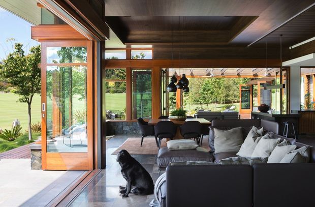 A contemporary open-plan living and dining area with extensive timber-framed glazing and open sliding doors connecting directly to a landscaped lawn and garden. The room features a large dark sectional sofa, lounge chairs, a dining table, pendant lighting, and dark timber ceiling lining and cabinetry. The polished concrete floor and warm timber frames create a strong indoor-outdoor connection, with a black dog in the foreground.