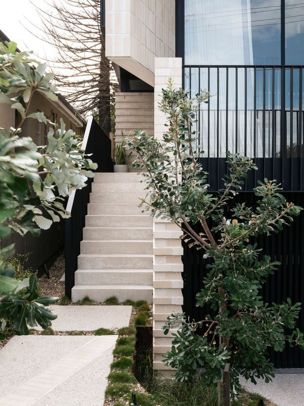 A contemporary residential exterior viewed from a landscaped front path, featuring a narrow concrete stairway rising between pale brick masonry walls and dark vertical metal fencing. The facade combines light textured cladding, large floor-to-ceiling glazed panels with black framing, and a projecting upper volume. Mature and newly planted greenery softens the entry sequence.