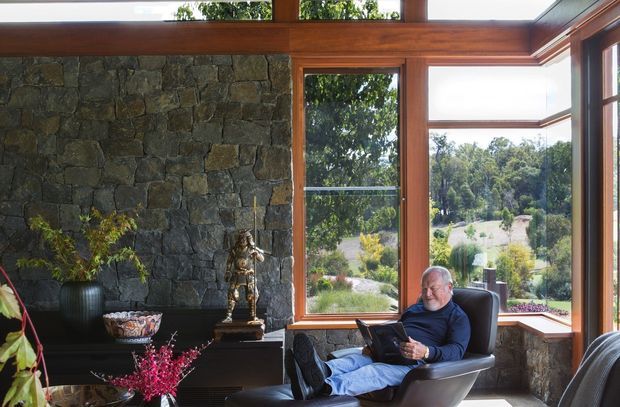 A sunlit interior sitting area with a large rough-stone feature wall, dark built-in joinery, extensive timber-framed glazing, and a seated person in a leather lounge chair reading. The windows overlook landscaped grounds, while a broad timber beam and clerestory-like glazing emphasize the room's connection to the outdoors. The space has a warm, natural, refined character.