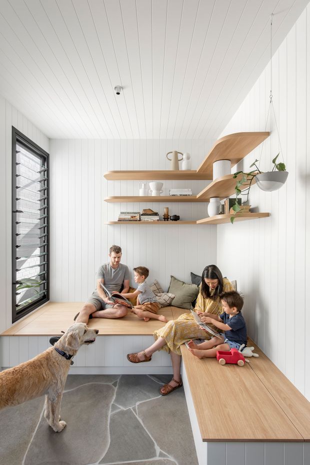 Bright, compact family living nook with white vertical wall and ceiling panelling, built-in timber window seating and a long timber bench, layered floating timber shelves, black louvred window, hanging planter, and a family gathered reading together. The space has a light, functional, contemporary feel with soft neutral furnishings and grey patterned flooring.