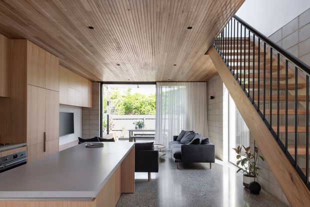 Open-plan contemporary living space with a kitchen island in the foreground, integrated timber cabinetry and television wall, dining area, and a compact lounge beyond. A timber-lined ceiling continues over the room, while a timber-and-black-metal staircase rises along the right side beside pale masonry walls. Full-height glazing provides abundant daylight and views to a planted outdoor area.