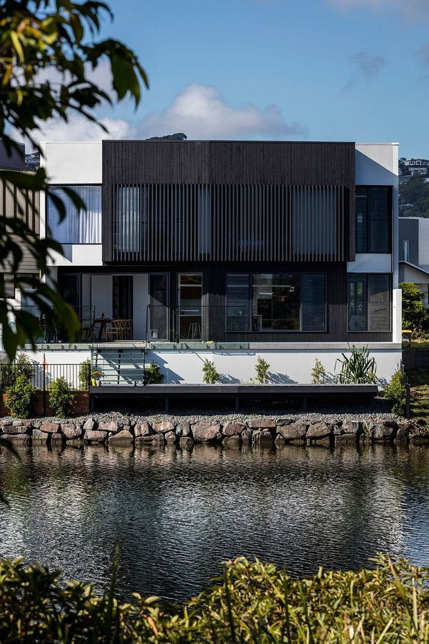Exterior view of a contemporary multi-storey waterfront residence across a narrow body of water. The house has a crisp white rendered facade, large dark-framed windows, and a prominent upper-level black vertical timber-look screen facade. A terrace and lower retaining edge sit above a rocky shoreline, with landscaped planting in the foreground.