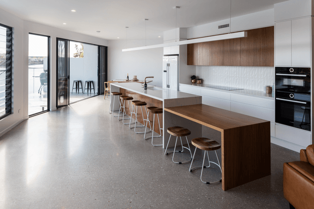 A bright, spacious contemporary kitchen with a large white island and integrated timber breakfast bar, seating for several people on stools. The room features white cabinetry, a warm timber feature panel, textured white splashback, integrated black ovens, pendant lighting, polished concrete flooring, and expansive black-framed glazing opening toward an outdoor area.