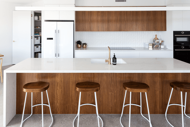 A bright contemporary kitchen dominated by a large white island with an integrated sink and brass-toned tap. The island front and overhead cabinetry feature warm mid-tone timber, complemented by white cabinetry, a white refrigerator, built-in black ovens, and a pale terrazzo-like floor. Four timber-topped stools with white metal frames line the island.