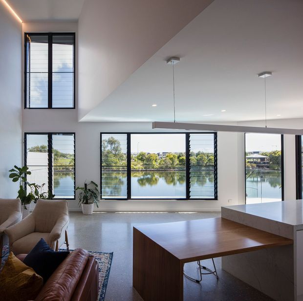 Bright, open-plan contemporary interior overlooking a calm waterway through expansive black-framed windows. The space combines a living area with leather and upholstered seating, indoor plants, and a timber dining table, alongside a large white kitchen island. A sloped ceiling, recessed lighting, and pendant fixtures create a clean, airy feel, while polished concrete flooring and warm timber furniture add contrast.
