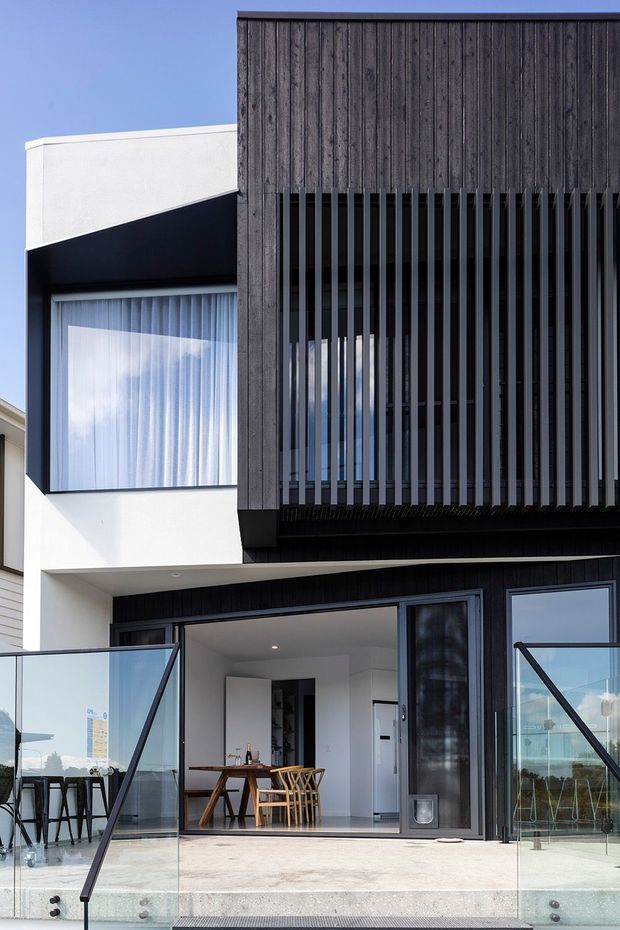 Contemporary multi-storey residential facade with crisp white rendered walls, dark vertical timber cladding and a prominent screened upper-level balcony or window enclosure. The ground floor opens onto a terrace through large glazed doors, with frameless glass balustrades, outdoor bar seating and a glimpse of a compact dining area inside. Strong geometric lines and contrasting black, white and timber finishes define the design.